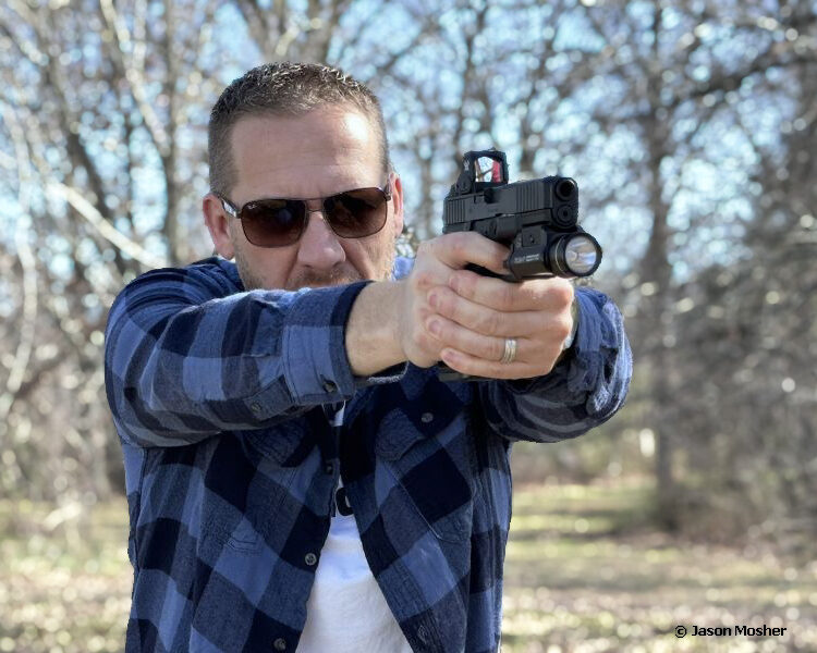 A person in a blue flannel shirt with black sunglasses shooting a handgun with a red dot mounted to it..