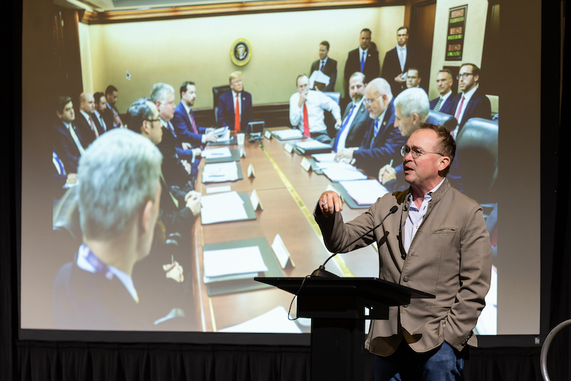 Mick Mulvaney speaking in front of photo from White House