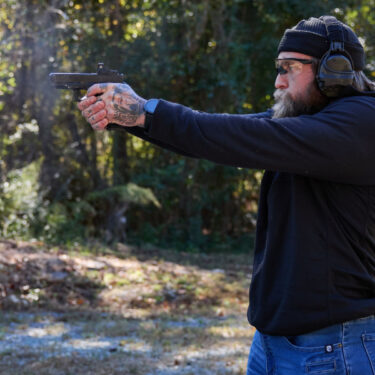 Blake Cook aims a handgun. He is wearing Liberator headset and Safariland range bag is in the background.
