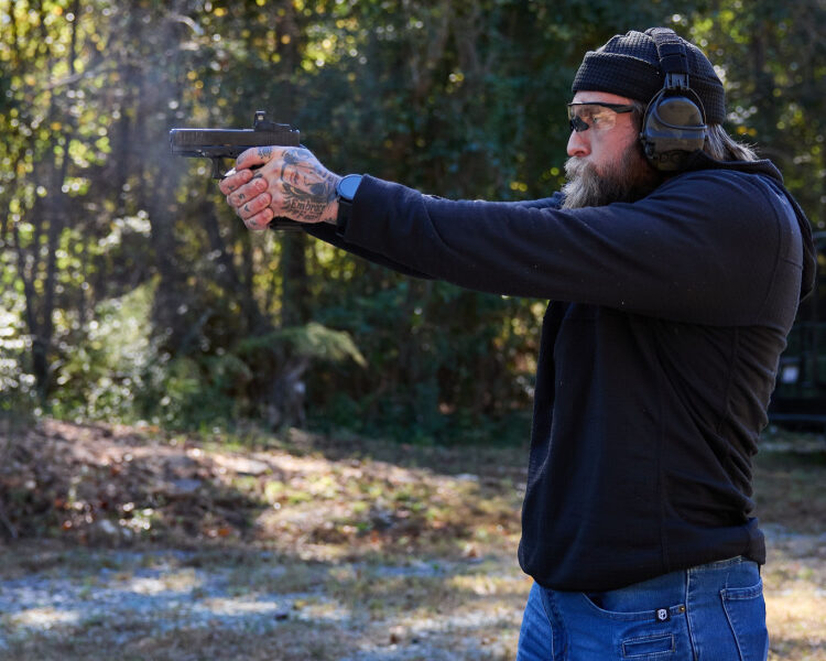 Blake Cook aims a handgun. He is wearing Liberator headset and Safariland range bag is in the background.