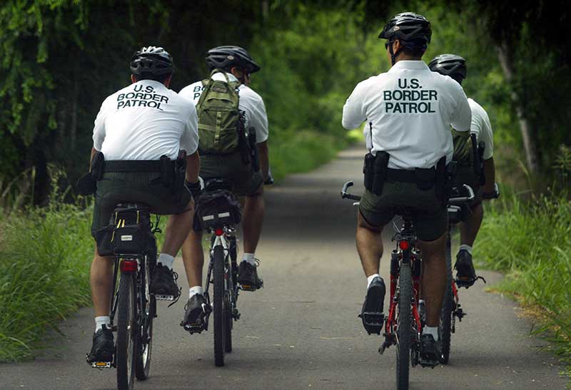 US Border Patrol agents riding bikes, two are wearing camelbak hands-free hydration packs.