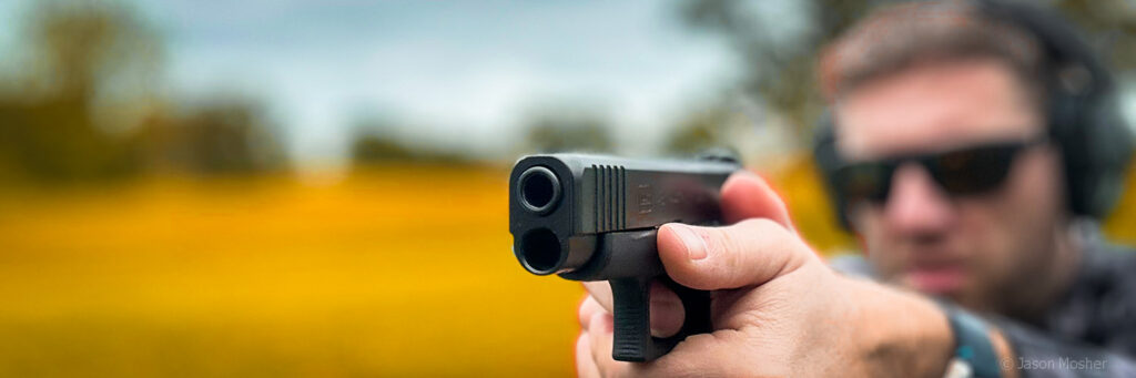 closeup view of the muzzle end of a handgun in hands, the man aiming it in the background is blurry, but he's wearing sunglasses and hearing protection earmuffs