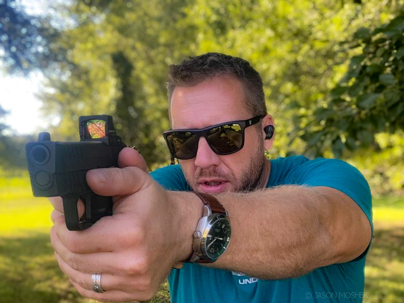 A person shooting a handgun in a field with a tree behind them with green leaves. The person is wearing a teal colored shirt and wearing a pair of sunglasses. 