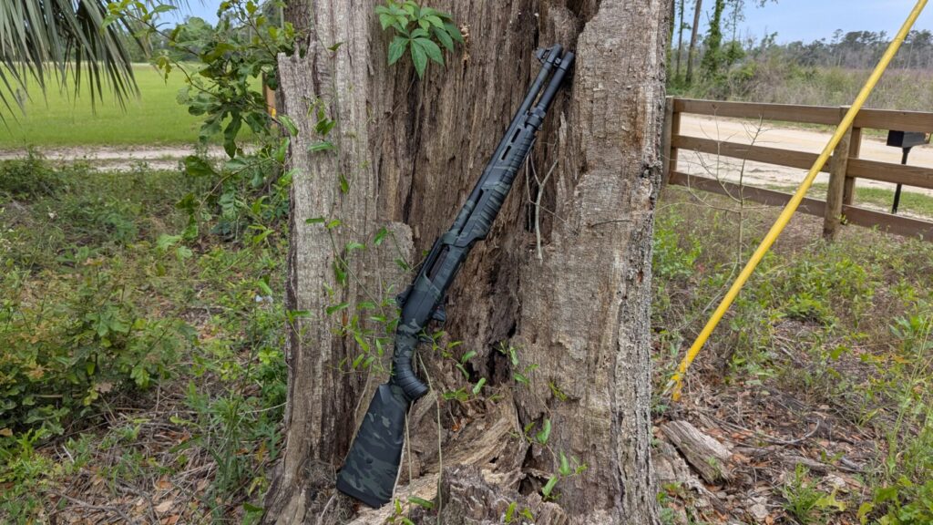 Nova 3 tactical shotgun leaning against a tree trunk. Wooden fence, gravel road, and fields are in the background.