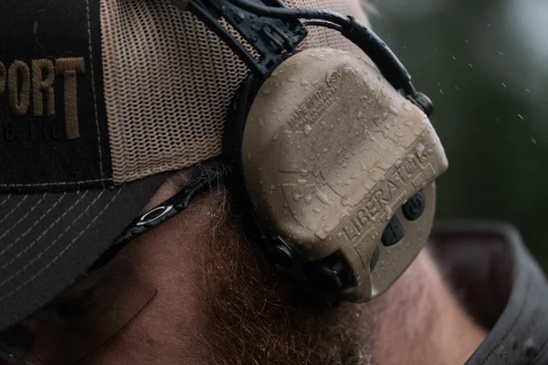 Closeup of Liberator HP hearing protection, on a bearded man wearing a ball cap and protective eyewear. It's raining, and you can see the rain drops on the Liberators.