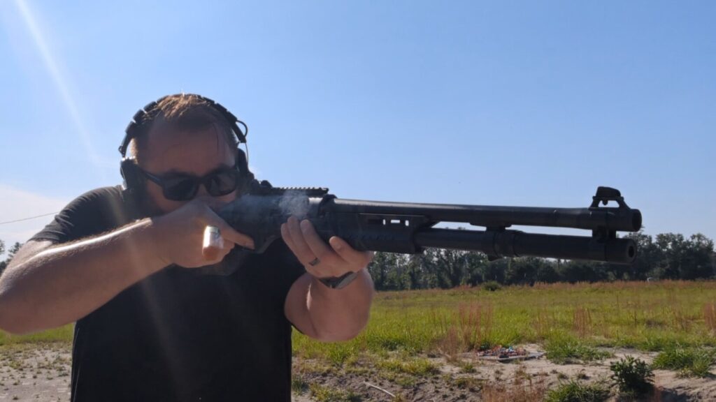 man wearing a black tshirt, sunglasses, and earmuff hearing protection shooting nova 3 pump action shotgun outdoors. Spent shotshell has just been ejected and is still in the air.