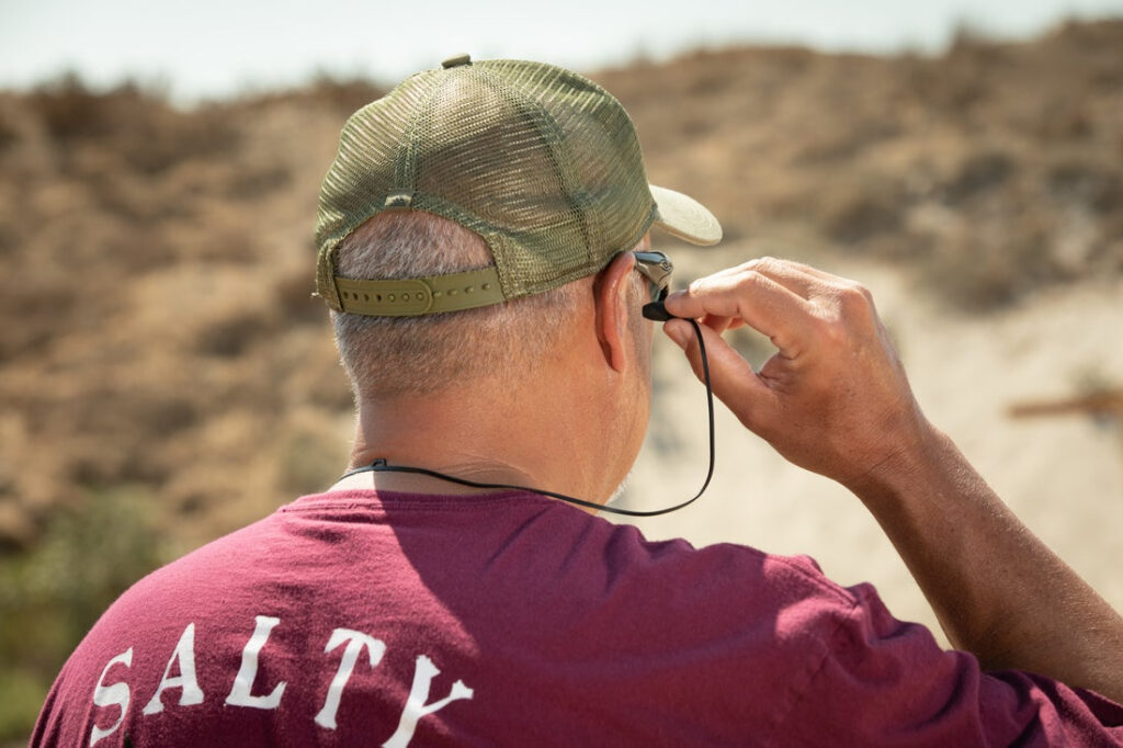 Rear view of man inserting Safariland Foam Impulse Hearing Protection into his ears. He's wearing a burgundy shirt with the word "SALTY" in white letters across the back. 