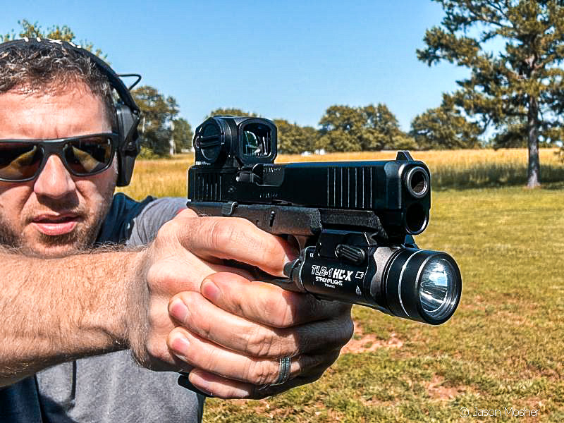 A person in a grey shirt and sunglasses, shooting a black handgun in a field. 