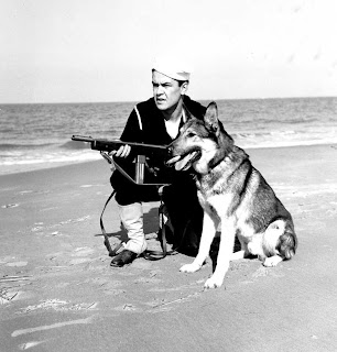This is a black and white photo from World War II showing a coastguardsman with a submachine gun kneeling with his german shepherd sentry dog
