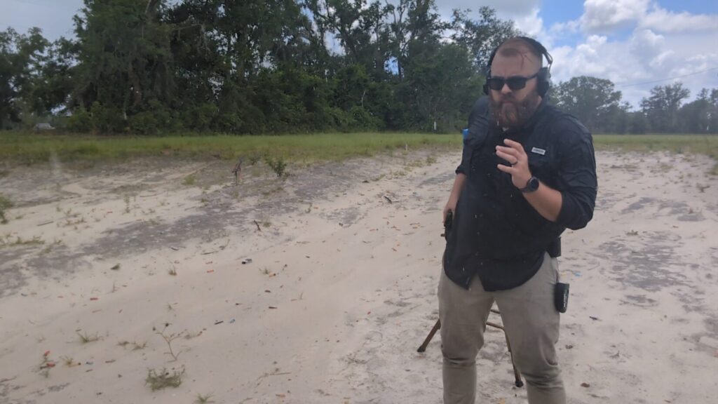 A man wearing ear protection and sunglasses draws a handgun from a holster at an outdoor range.
