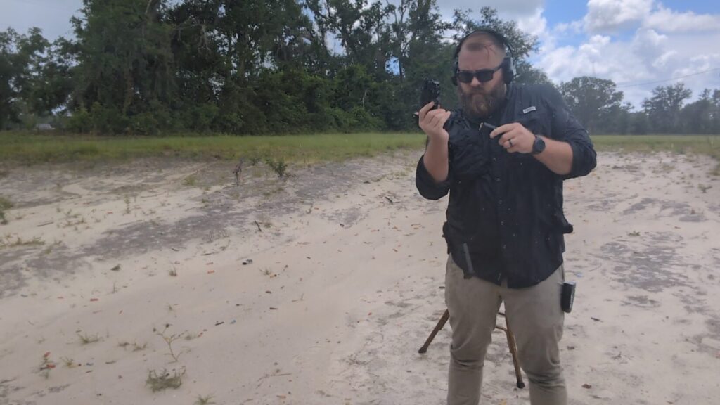 A man wearing ear protection and sunglasses performs a reload with a handgun and magazine at a sandy outdoor range.