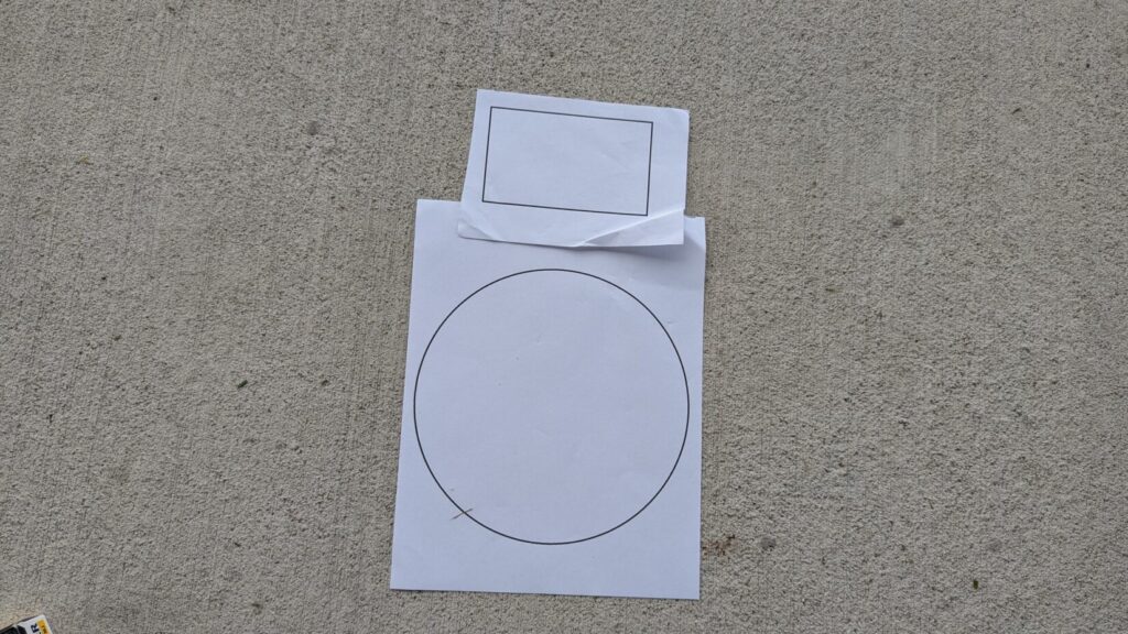 A paper target with a circle and rectangle drawn on it, lying on concrete.