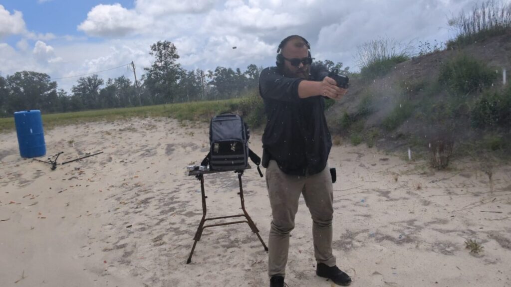 A man wearing ear protection and sunglasses holds a handgun with an optic, aiming forward at an outdoor range.