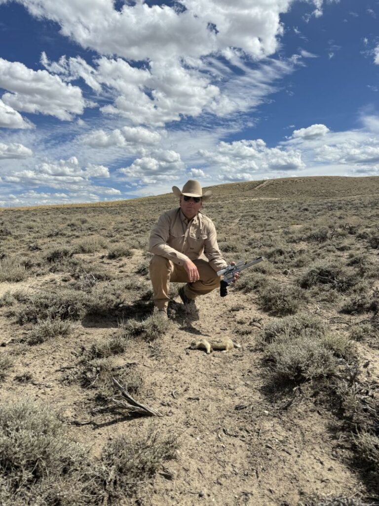 man crouched down with a Ruger super redhawk in .22 hornet topped with a 4x scope, prairie dog on the ground in front of him.