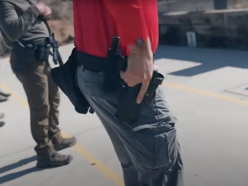 A student on the range wearing a red shirt, grey pants and ear muffs, draws his handgun from his holster just after firing his rifle at a target. 