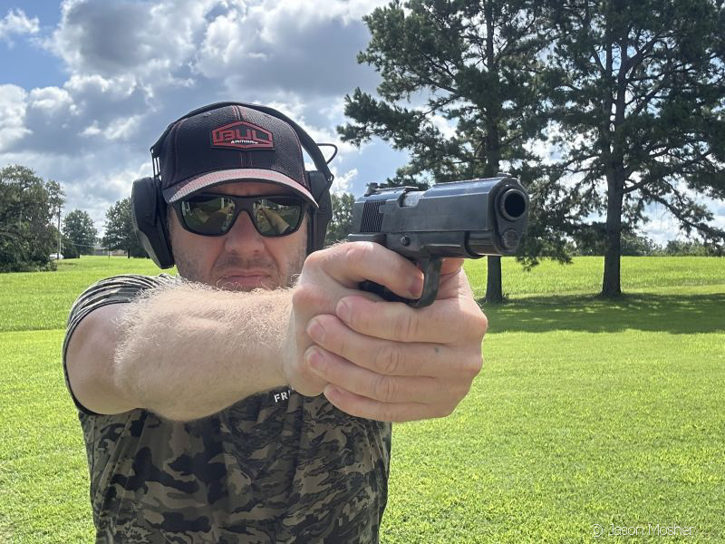 man aiming a Llama .45 ACP 1911-style handgun. He's wearing a camo tshirt, a Bul Armory hat, and hearing protection muffs.