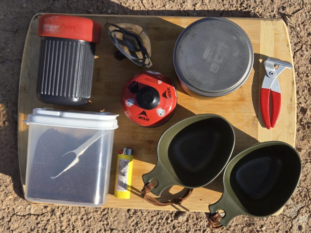 Coffee kit on a a cutting board that includes an aero press, fuel container, stove, pot lifter, pot, and two cups.