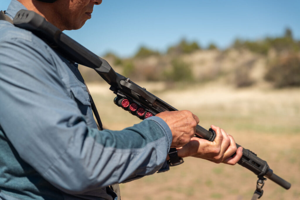 A shotgunner sustain loads the magazine tube of a shotgun with the firearm over his shoulder