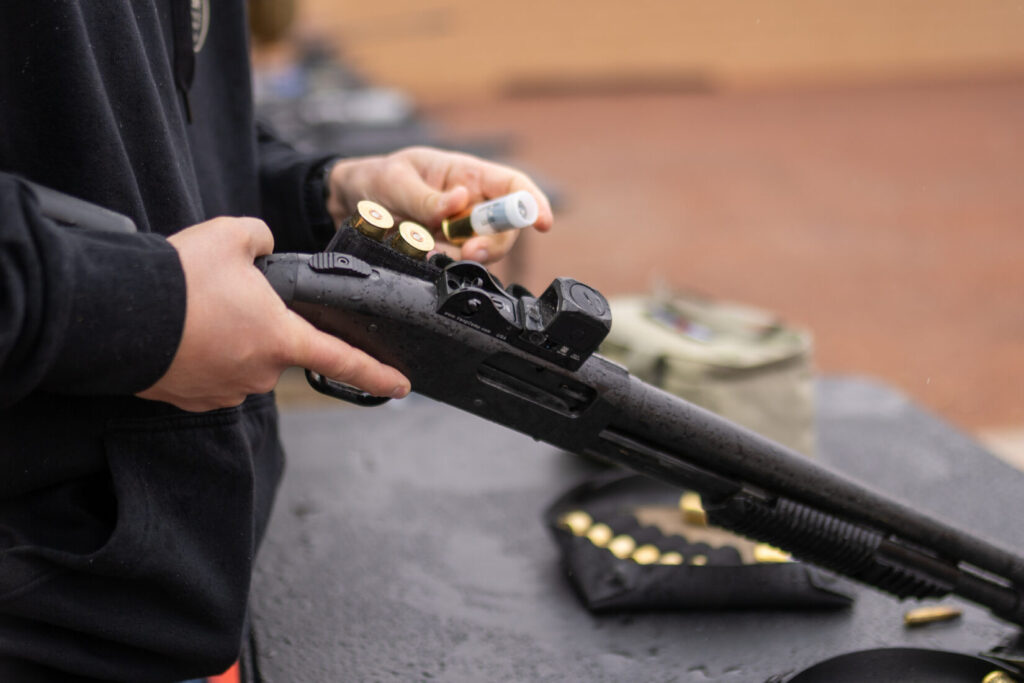 A 12 gauge slug is held in the left hand of a shotgunner about to load it into the Mossberg vang comp standard 590