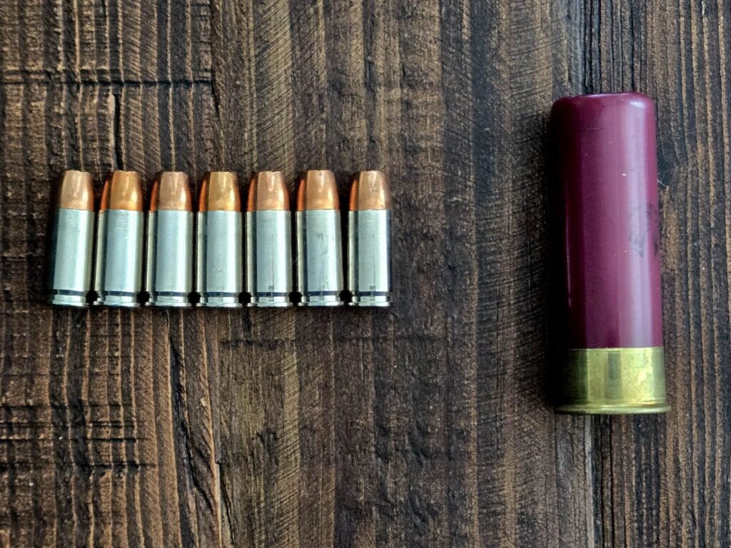 seven 9mm hollow point bullets next to a single buckshot 12 gauge shell on a wooden backdrop.