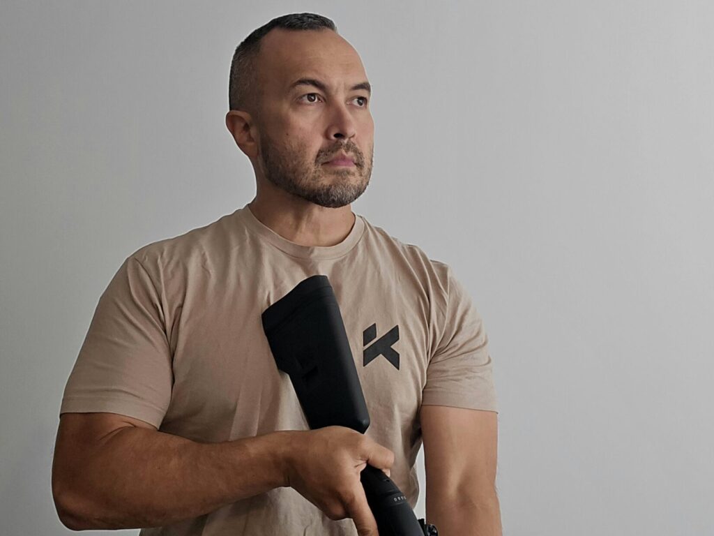 A man in a tan shirt holds a shotgun with the buttstock mounted underneath his dominant eye against his chest.