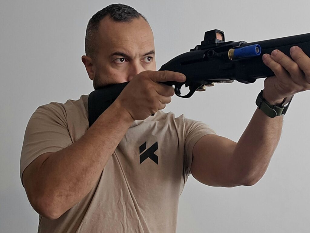 A man in a tan shirt with a Beretta 1301 shotgun takes aim through a Holosun 509T optic.