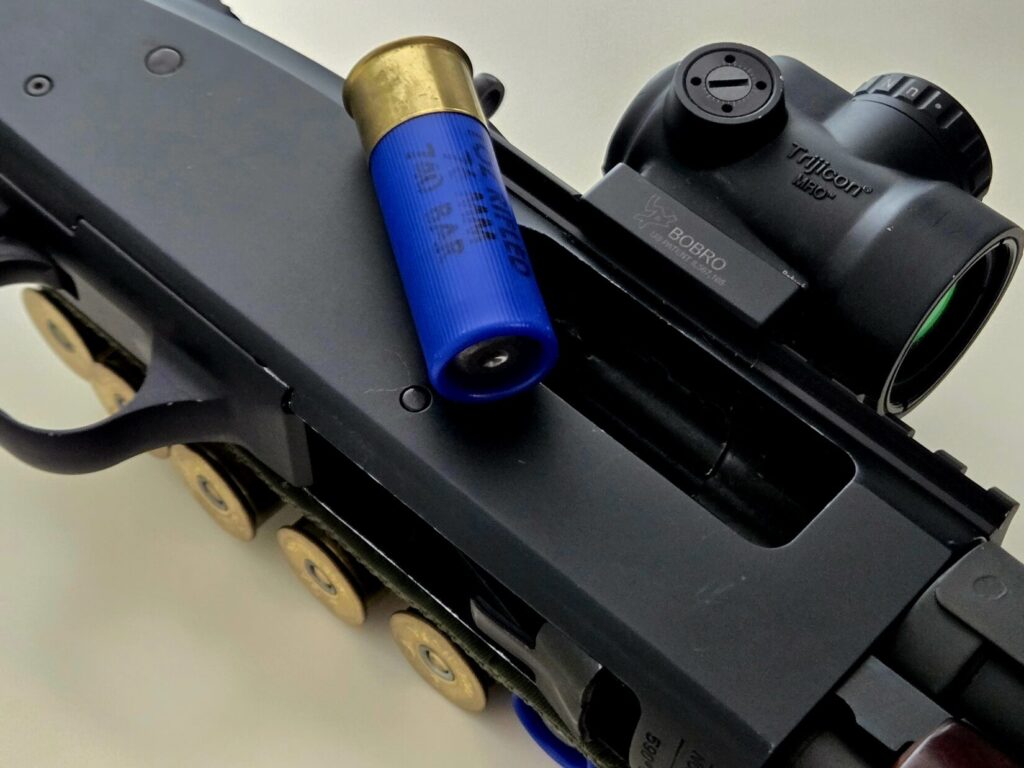 A blue 12 gauge slug sits on the receiver of a 12 gauge shotgun with a Trijicon MRO Optic mounted in a bobro throw lever mount.