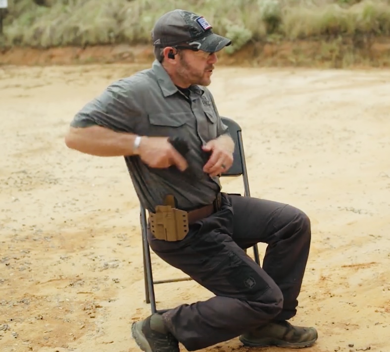 Rick Hogg of War HOGG Tactical is seated in a chair at an outdoor range, drawing a handgun from an OWB holster. He's demonstrating how to use your feet in assistance to making a pivot move to be able to engage a target at an angle