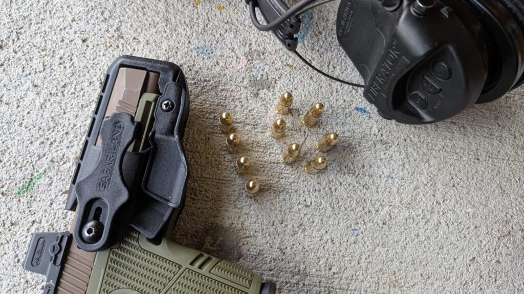 Pistol in Schema holster, loose brass cartridges, and ear protection on concrete.