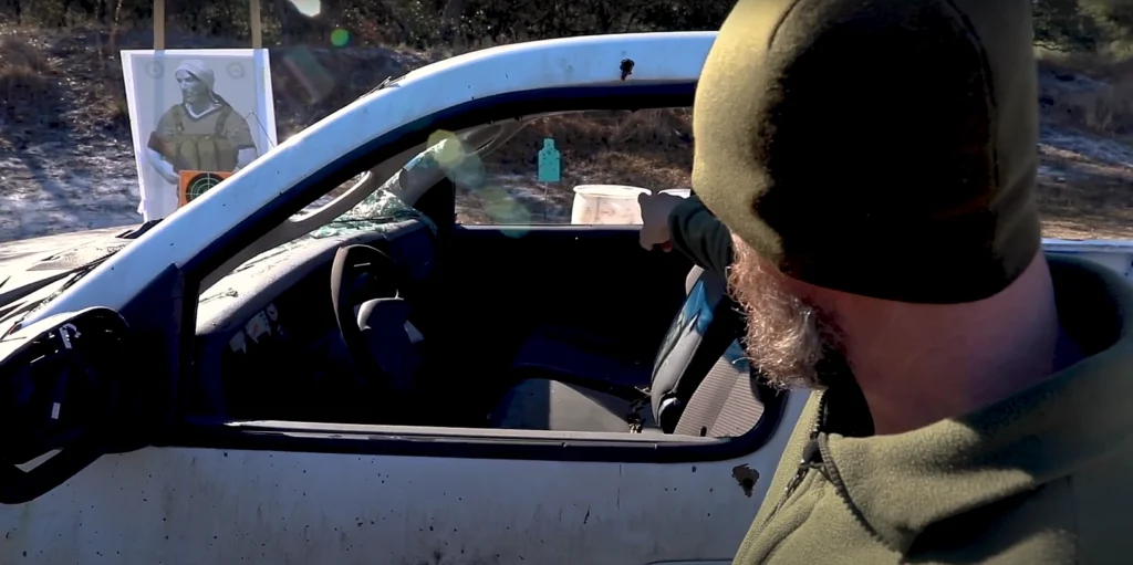 man demonstrating how a distant shooter has an advantage in a car attack. He's pointing through car windows at a blue target several yards on the other side of the car.