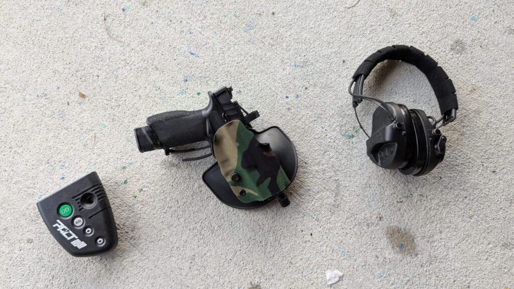 Black pistol in camo holster, dry fire device, and ear protection.