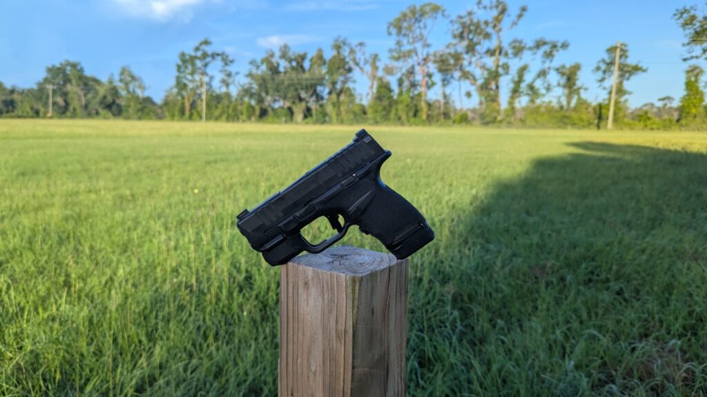 A black Hellcat pistol balanced on a wooden post in a green field under a blue sky, with a shadow visible.