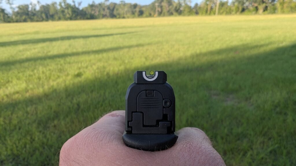 A first-person view of aiming a Hellcat pistol, showing the front sight with a bright green dot, over a blurred green field.