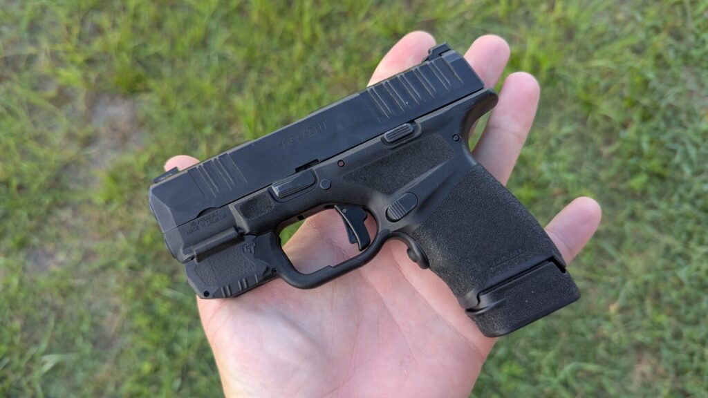 A black Hellcat pistol held in the palm of a hand, with a grassy background.