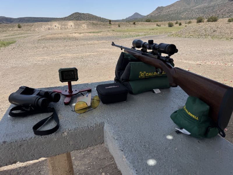 view of long-range shooting setup from behind the bench. Left to right, binoculars, Athlon Optics Rangecraft Velocity Pro Radar Chronograph (RVPRC), field notebook, sunglasses, soft box, scoped rifle, and Caldwell rifle rests. Mountains in the background.