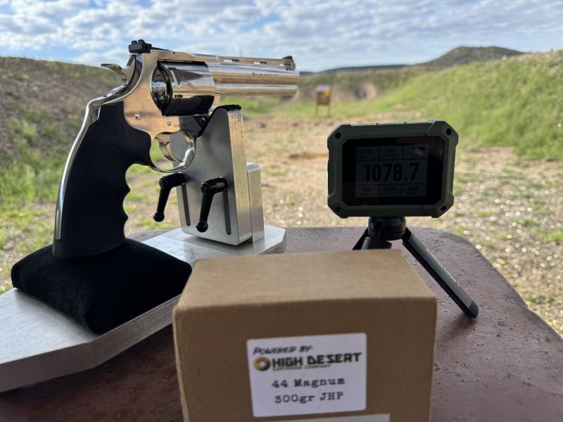 view from behind the bench at an outdoor range with mountains in the background. Revolver on a rest to the left, Rangecraft Velocity Pro Radar Chronograph to the right, box of .44 Magnum ammo from High Desert in the foreground.