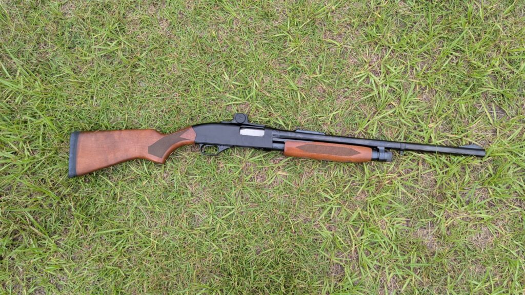 A shotgun with a red dot sight mounted, lying on green grass, viewed from above.