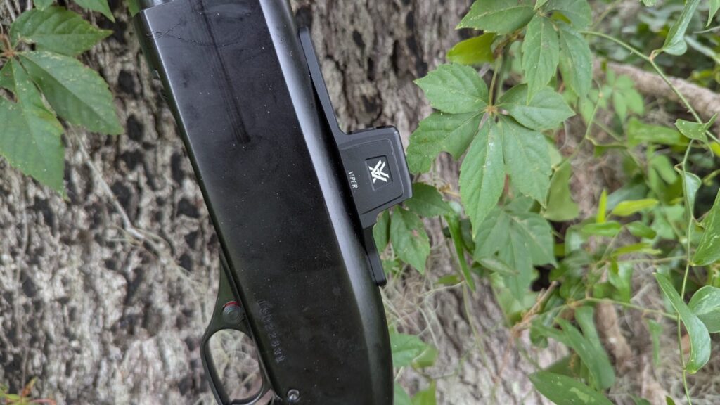 Side view of a shotgun with a Vortex Viper red dot sight, against a tree.