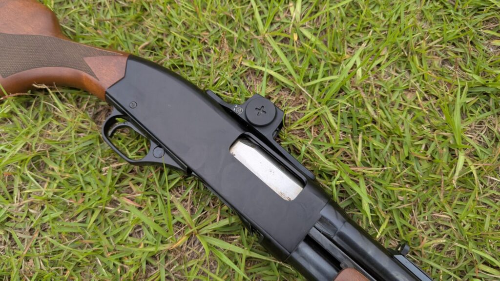 Angled top-down view of a shotgun with a red dot sight, lying on green grass.