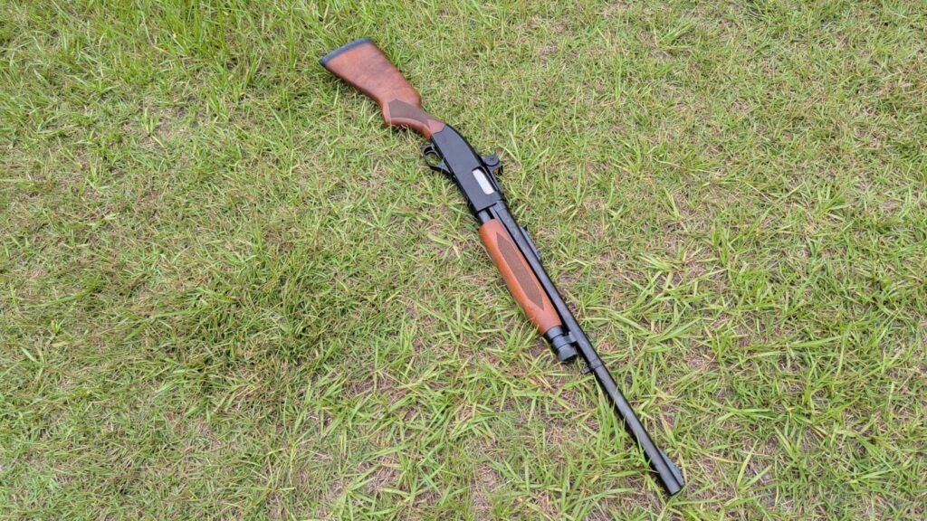 An entire shotgun with a red dot sight, lying diagonally on green grass, viewed from above.