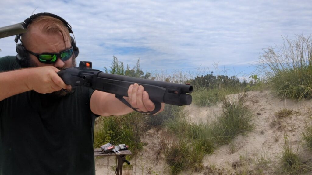 Man aiming shotgun with red dot sight, spent casing at outdoor range.
