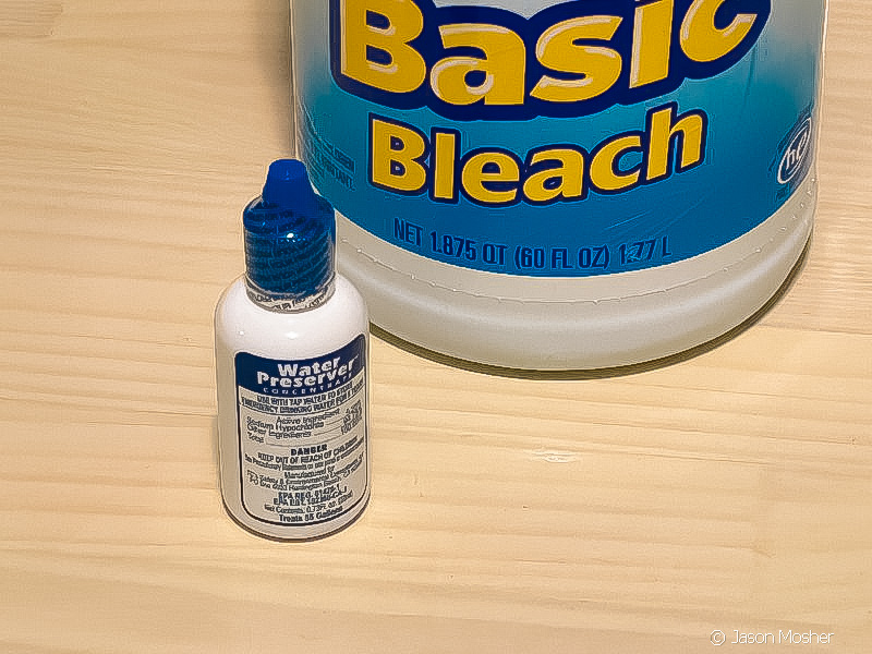 A small bottle with a blue lid containing water preserver, next to a bottl eof bleach on a wood counter.