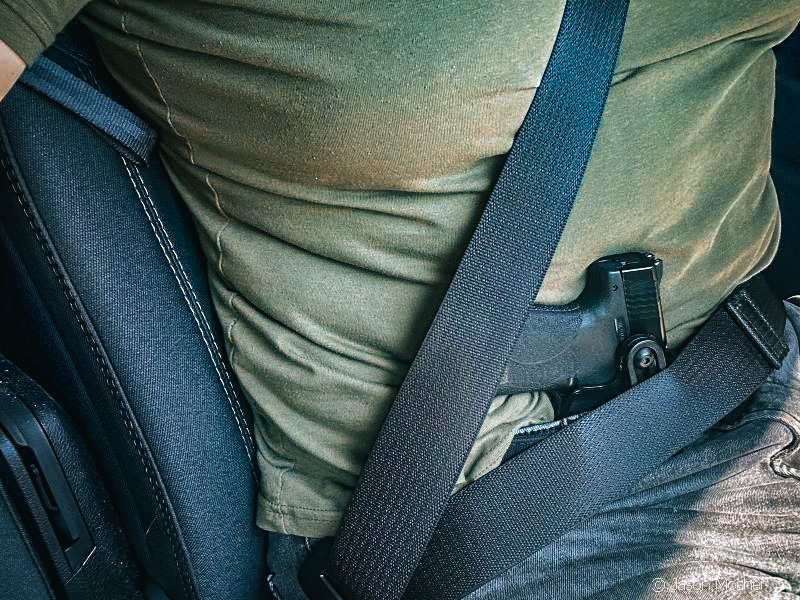 A person with a green shirt sitting in a truck with the seat belt on and a small black gun tucked in a IWB holster in the appendix position. The seat belt is covering up part of the gun. 