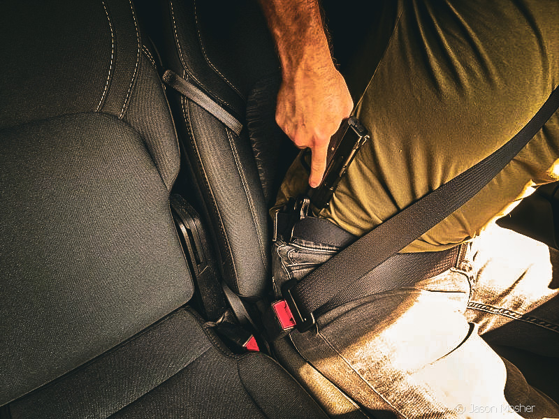 A person with a green shirt sitting in a truck with a firearm on, drawing a gun from a holster on their right side. 