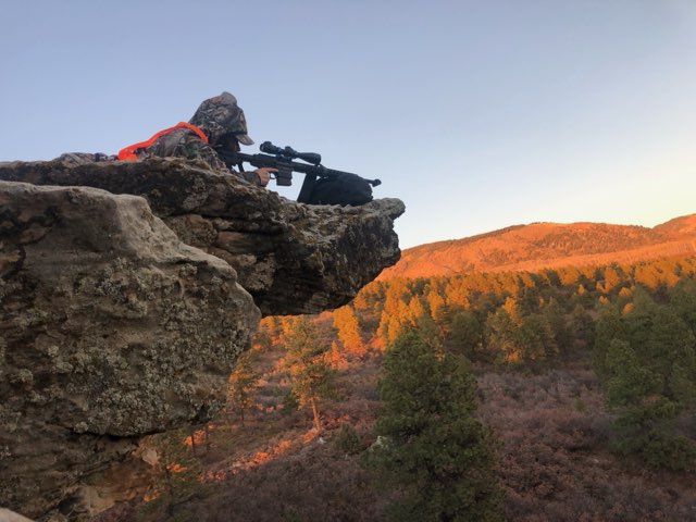 hunter in prone position on a rocky ledge, looking through a rifled scope