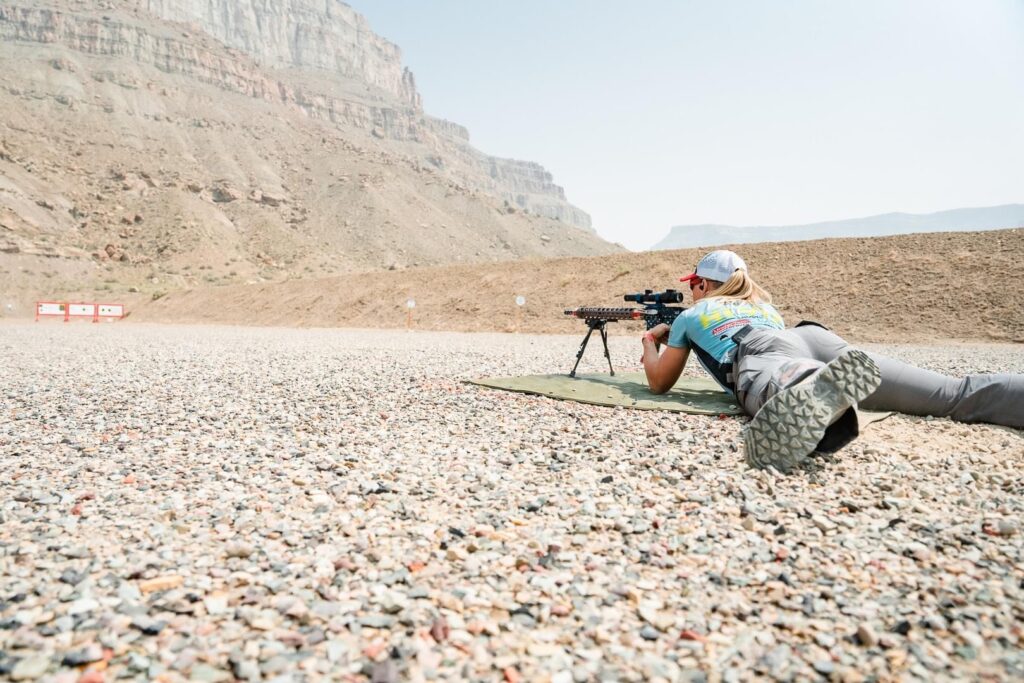 shooter in prone position looking through rifle scope aimed at target, mountain in the background