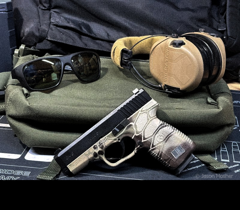 A Kahr, brown camo 9mm handgun leaned up against a green bag with a pair of brown Safariland Liberator earmuffs and black Vortex sunglasses in the background. 