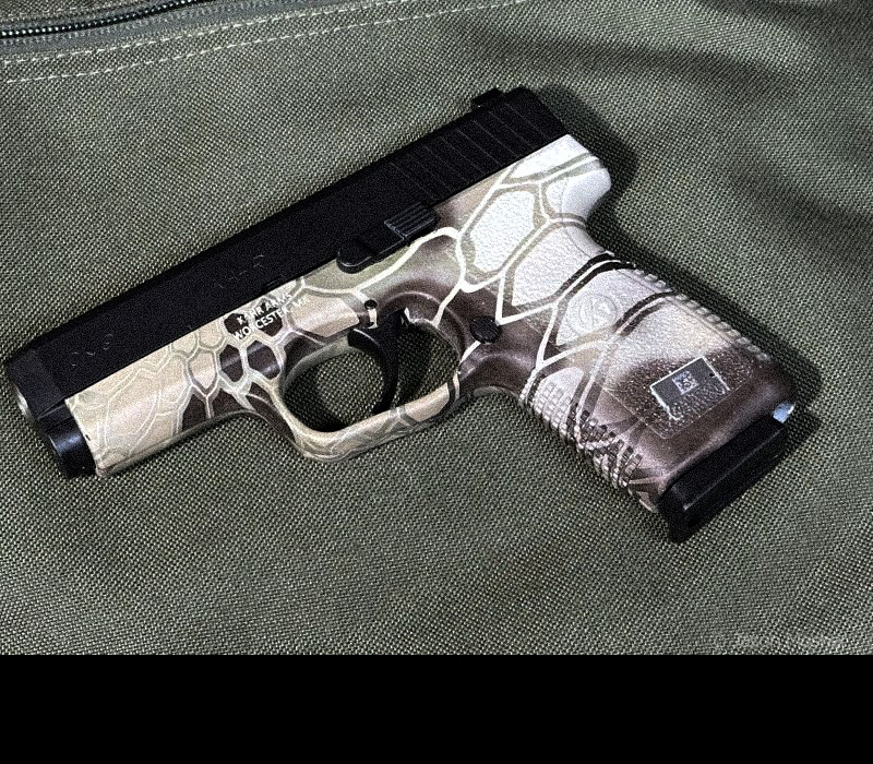 A brown camo colored Kahr SW9 handgun with a black slide, laying on a green background. 