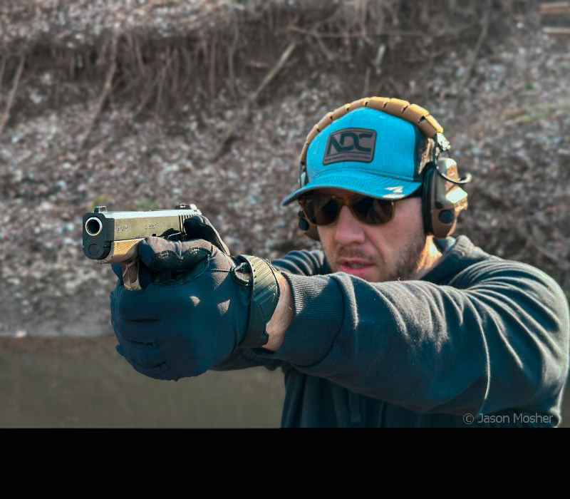 A person with a grey hat and brown sunglasses shooting the Kahr SW9. Brown grass and some water can be seen behind the person. 