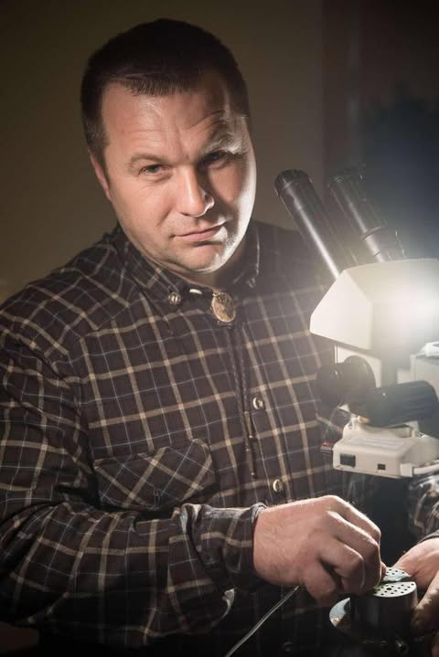 An engraver standing behind a microscope wearing a plaid shirt and a bolo tie